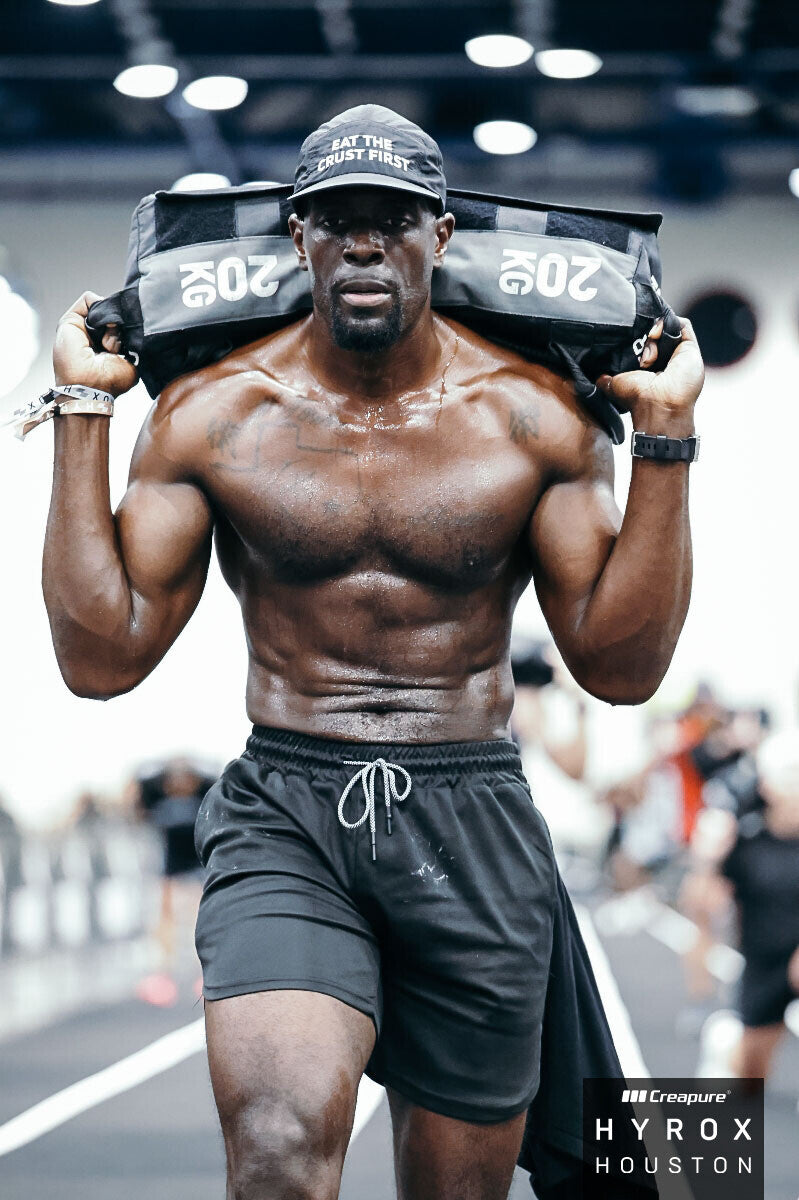 Joshua carrying a sandbag at HYROX Houston while wearing his black ETCF performance hat